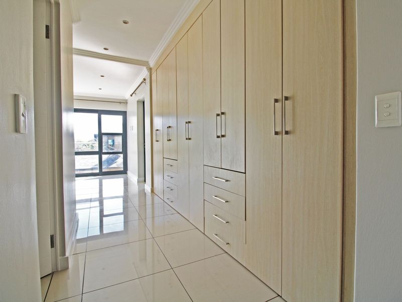 A bright, modern hallway featuring extensive built-in cabinetry with light wood finish, polished tiled flooring, and recessed lighting leading to a glass sliding door.