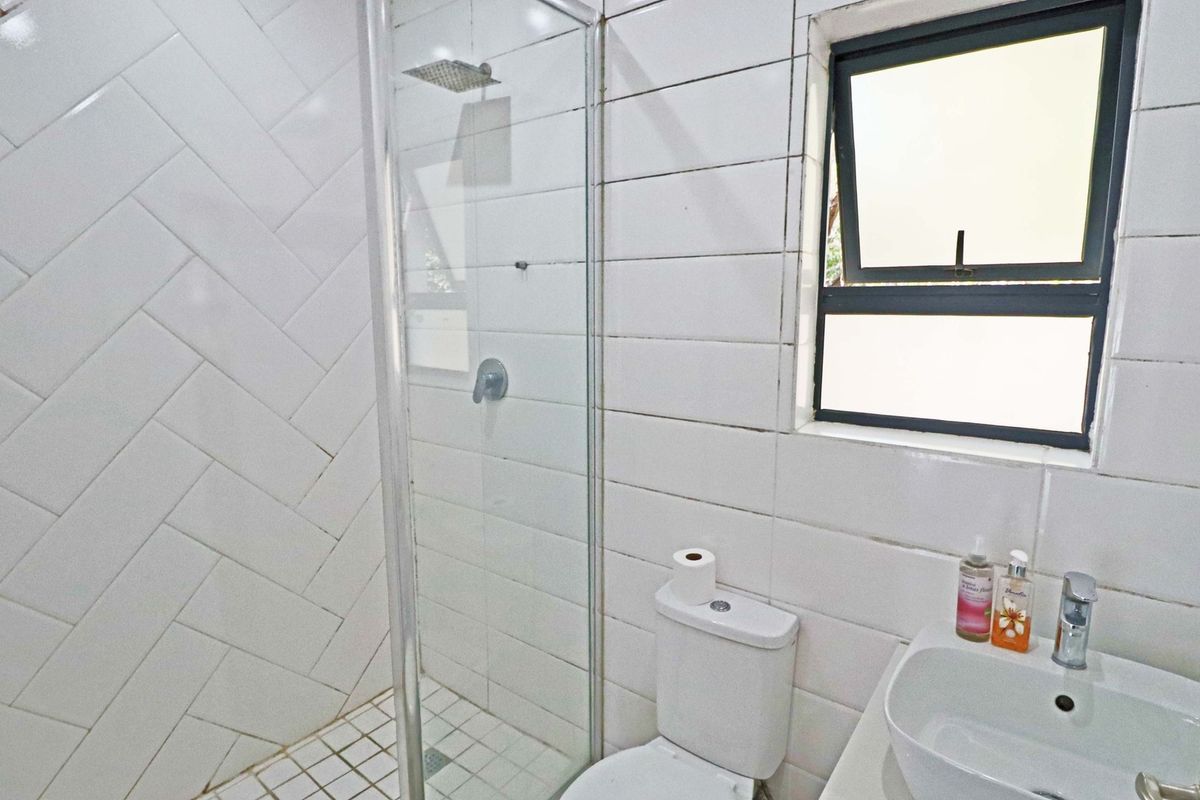 A functional bathroom featuring a glass-enclosed shower with herringbone wall tiling, a standard toilet, and a wall-mounted basin with a window for natural light.