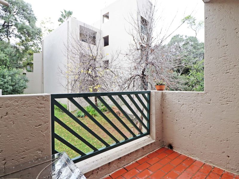 A private balcony featuring terracotta tile flooring and a metal balustrade, overlooking the apartment complex grounds.