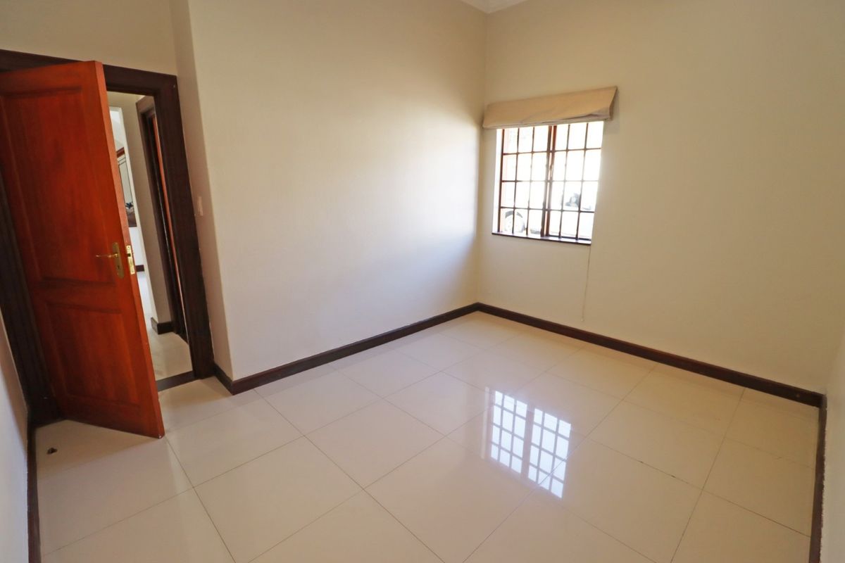 A bright, empty bedroom featuring neutral tiled flooring, a timber door, and a window fitted with security bars.