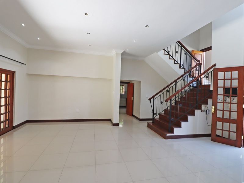 Spacious, light-filled living area featuring neutral tiled flooring, recessed lighting, and a classic wooden staircase with decorative wrought iron railings.