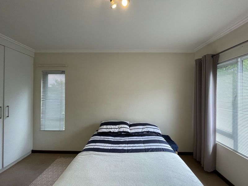 A well-lit bedroom featuring neutral walls, carpeted flooring, a built-in wardrobe, and dual window treatments including venetian blinds and curtains.