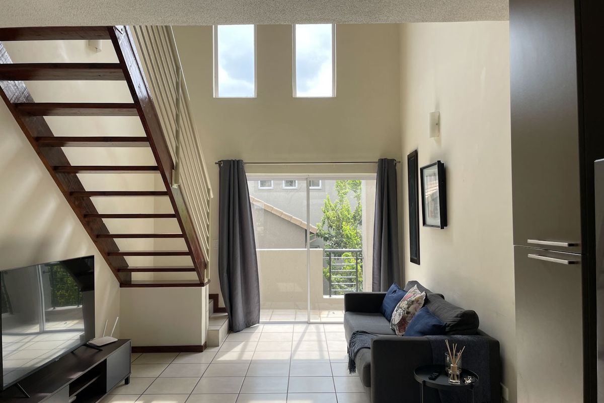 Spacious double-volume living area featuring tiled floors, a wooden staircase, and large sliding doors leading to a private balcony.