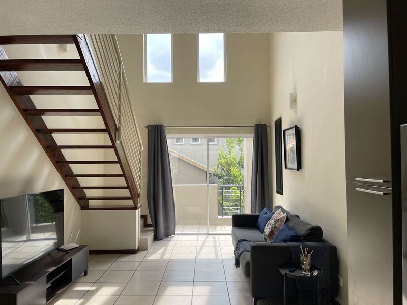 Spacious double-volume living area featuring tiled floors, a wooden staircase, and large sliding doors leading to a private balcony.