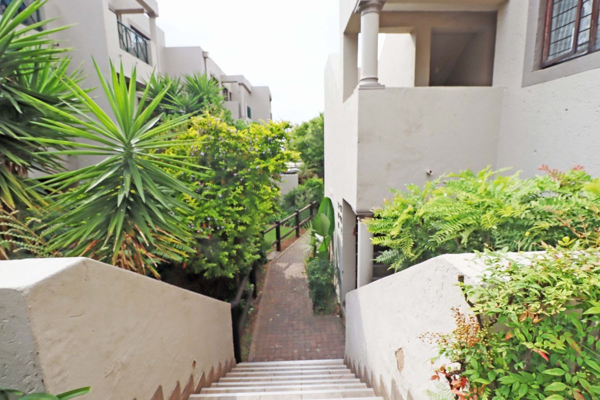 Exterior view of an apartment complex featuring a paved walkway, staircase, and balcony access surrounded by landscaped greenery.