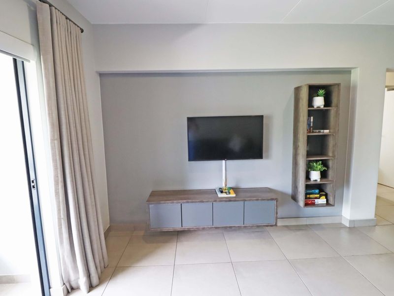 A modern living room featuring neutral tiled flooring, a wall-mounted media console, and integrated shelving, with access to an outdoor area via sliding doors.