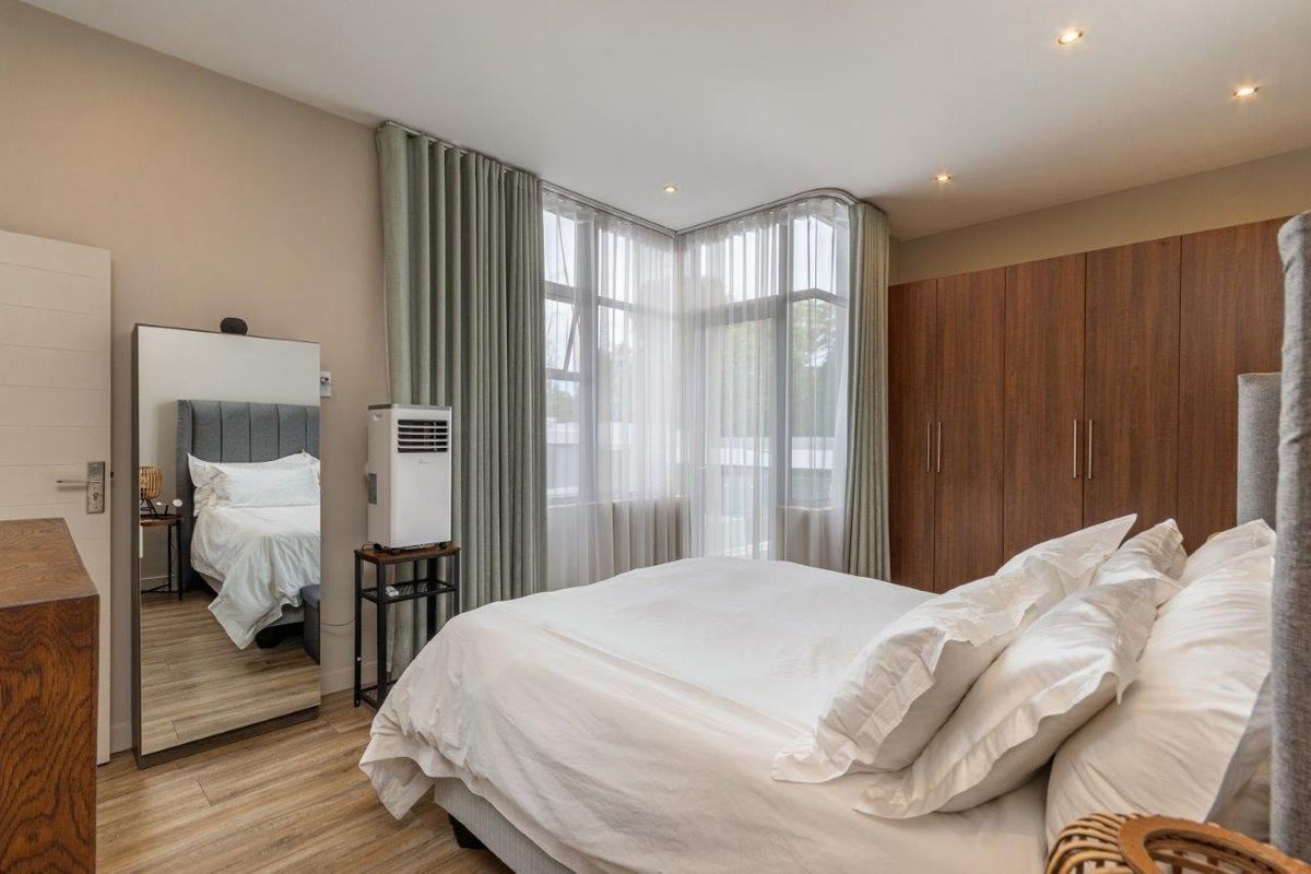 A well-lit bedroom featuring built-in wooden cupboards, modern laminate flooring, and large corner windows with floor-to-ceiling curtains.