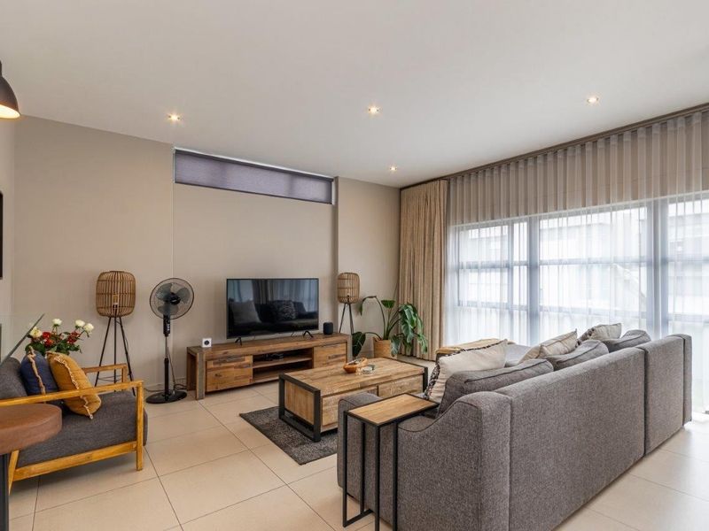 A well-lit, modern living room featuring neutral tiled flooring, recessed downlighting, and large windows with integrated curtain tracks.