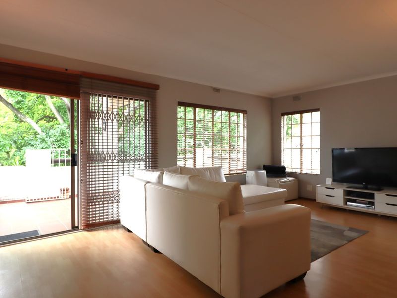 A bright living room featuring laminate flooring, large windows with timber blinds, and a sliding door providing access to an outdoor balcony area.