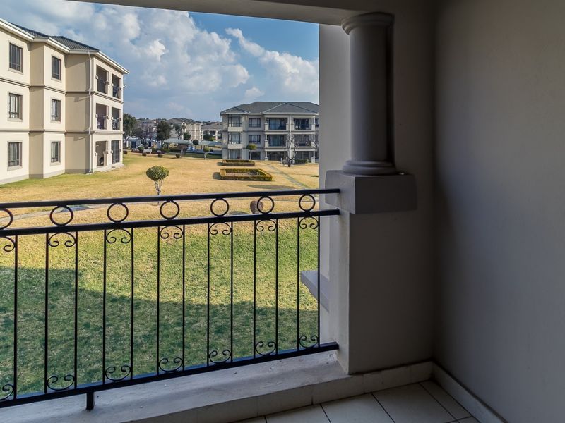 A covered balcony featuring a decorative wrought iron balustrade and tiled flooring, offering views over the apartment complex grounds.