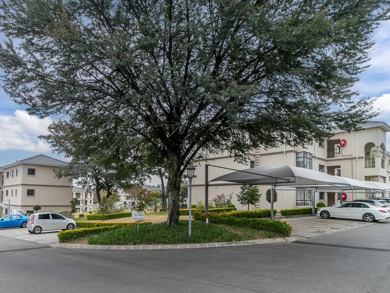 A well-maintained apartment complex exterior featuring paved driveways, covered parking bays, and established landscaped gardens.