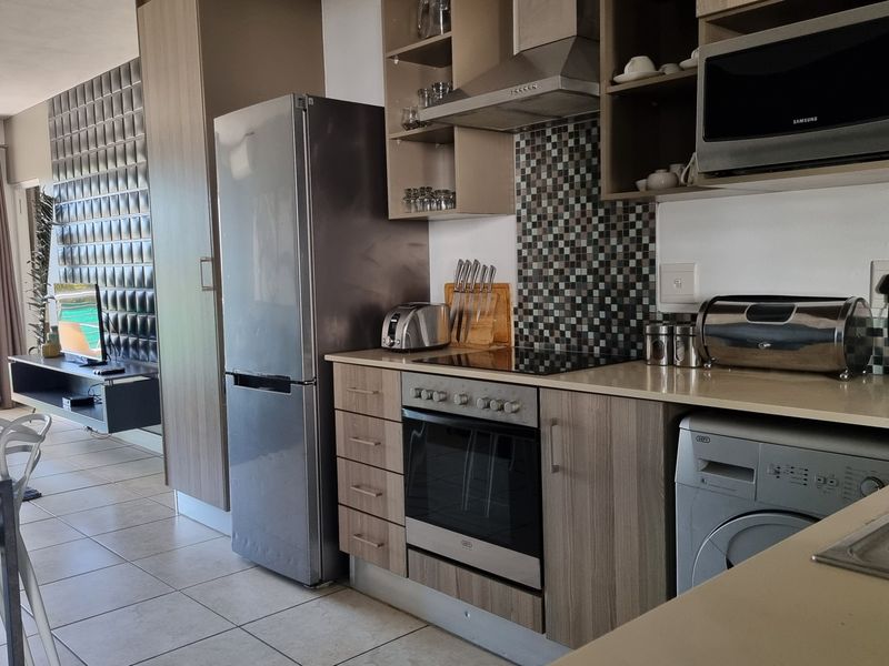 Modern open-plan kitchen featuring wood-finish cabinetry, mosaic backsplash, built-in appliances, and tiled flooring, seamlessly flowing into the living area with balcony access.