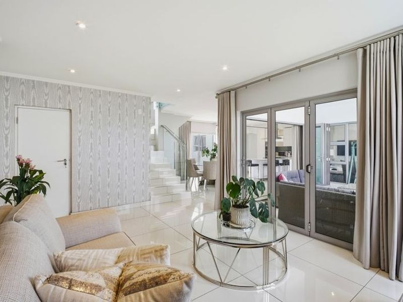 Bright open-plan living area featuring polished tiled floors, a modern glass balustrade staircase, and folding doors leading to an outdoor patio space.