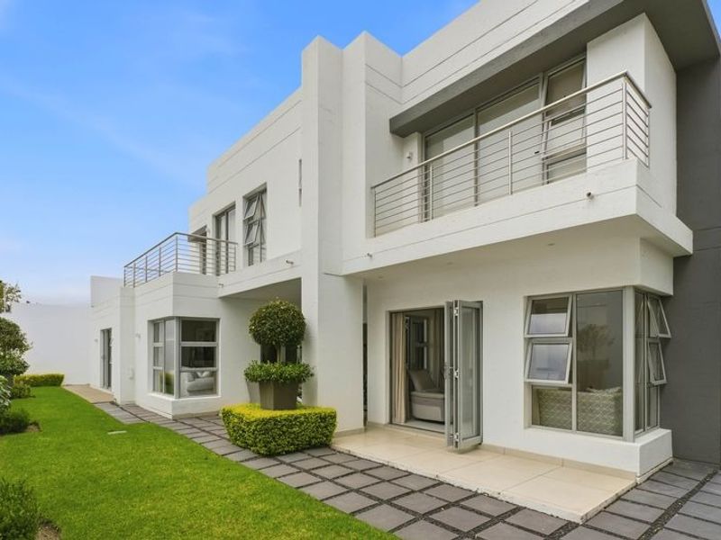 Modern double-storey cluster home featuring a clean white facade, upper-level balconies with stainless steel balustrades, and a well-maintained garden with paved pathways.