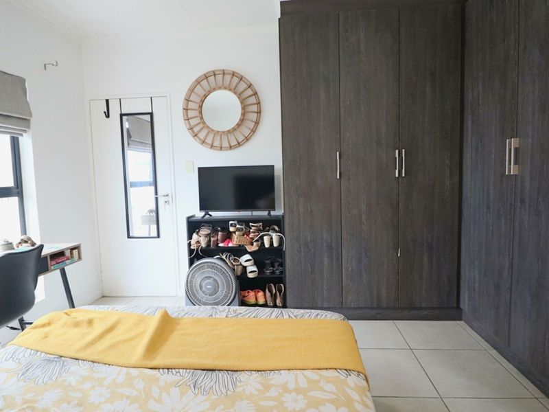 A well-lit bedroom featuring dark wood built-in cupboards, neutral tiled flooring, and a window with a fitted roller blind.