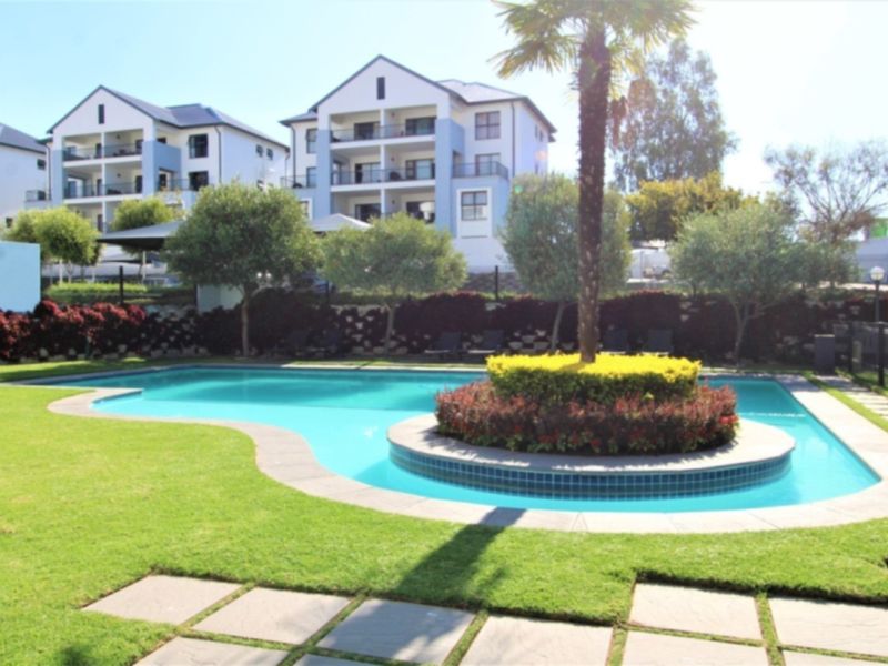 A well-maintained communal swimming pool area featuring landscaped gardens, paved surrounds, and a central palm tree island within an apartment complex.