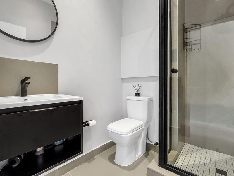 Modern bathroom featuring a sleek vanity with black basin mixer, round mirror, white toilet, and a glass-enclosed shower with tiled flooring.