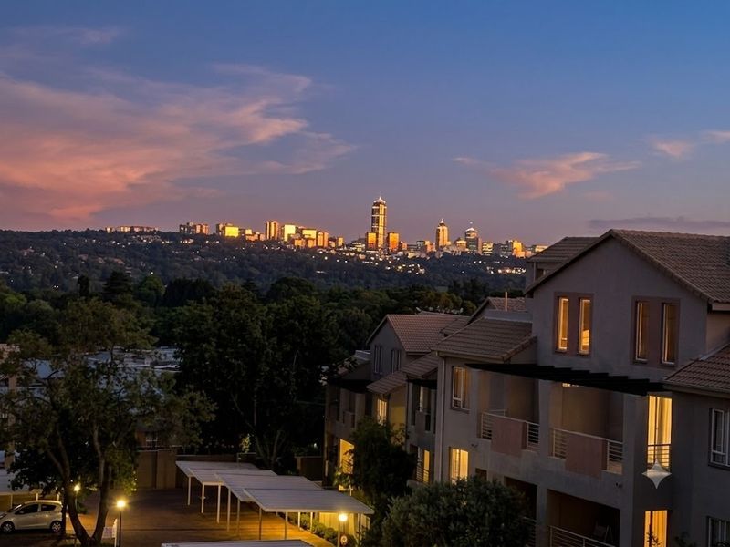 A scenic elevated view of the city skyline at dusk, overlooking the apartment complex with its tiled roofs and covered parking areas.