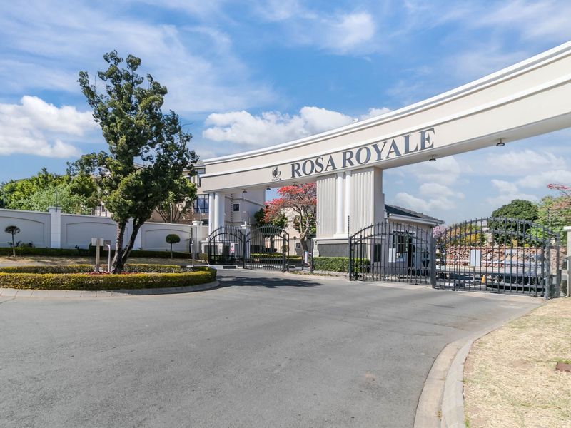 The main entrance gate and security access point for the Rosa Royale apartment complex, featuring a paved driveway and well-maintained perimeter landscaping.