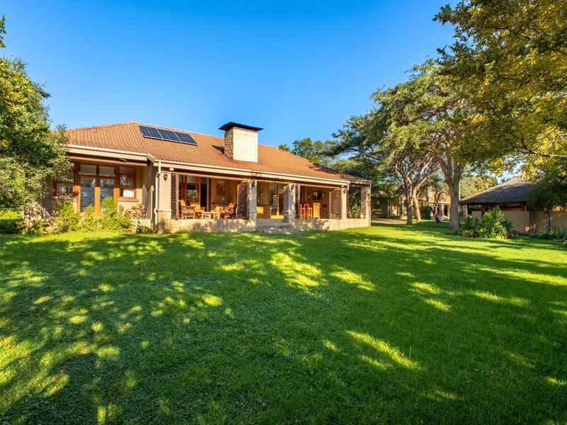 A spacious rear view of the house featuring a large manicured lawn, a covered patio area, and a stone chimney, set within a lush garden.