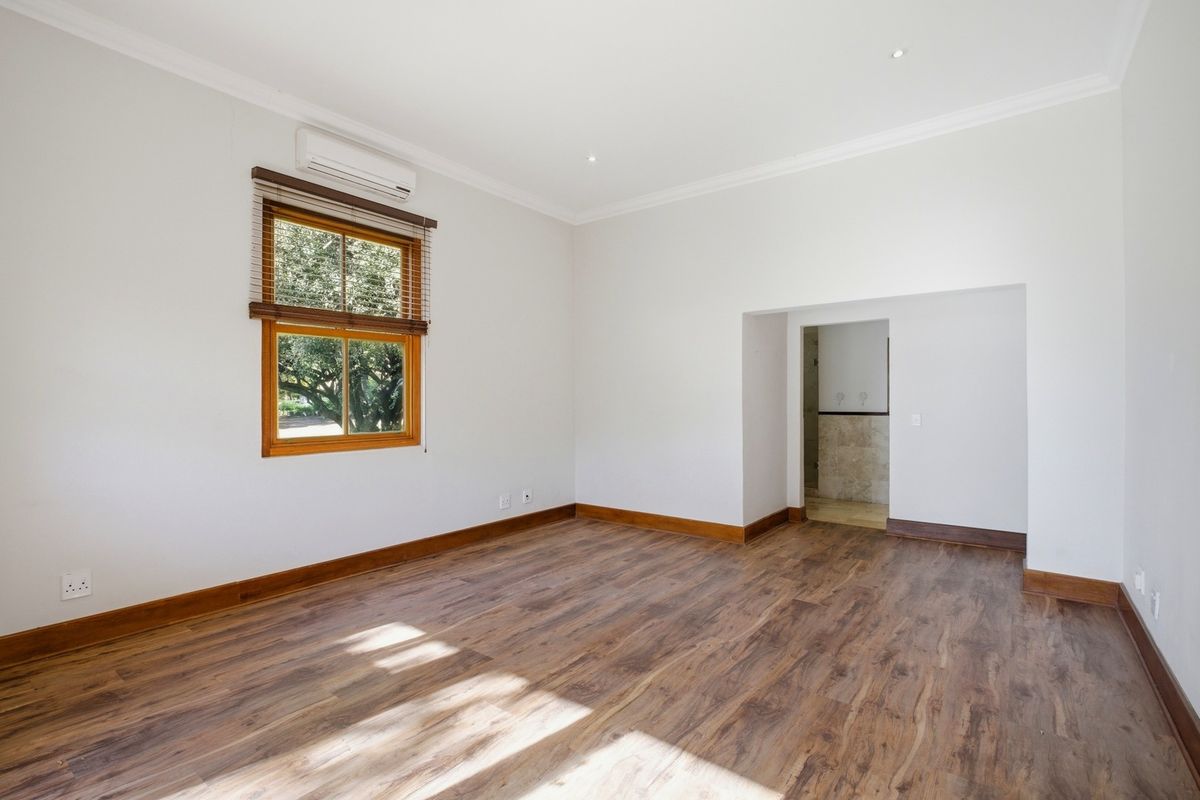 A bright, empty bedroom featuring laminate flooring, timber-framed windows, and a wall-mounted air conditioning unit, with an adjacent ensuite access point.