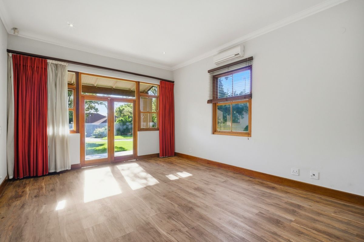 A bright, empty living room featuring laminate flooring, timber-framed windows and doors, and a wall-mounted air conditioning unit with direct access to the garden.