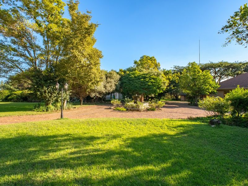 A spacious, well-maintained garden featuring a brick-paved driveway, a central stone fountain, and mature trees set within a lush green lawn.