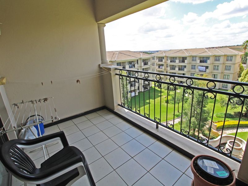 A covered balcony featuring tiled flooring and a decorative wrought iron balustrade, overlooking the apartment complex grounds.