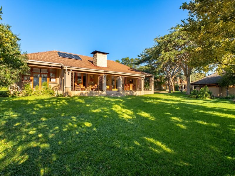 A well-maintained rear exterior view of the residence featuring a spacious lawn, covered patio, stone chimney, and roof-mounted solar panels.