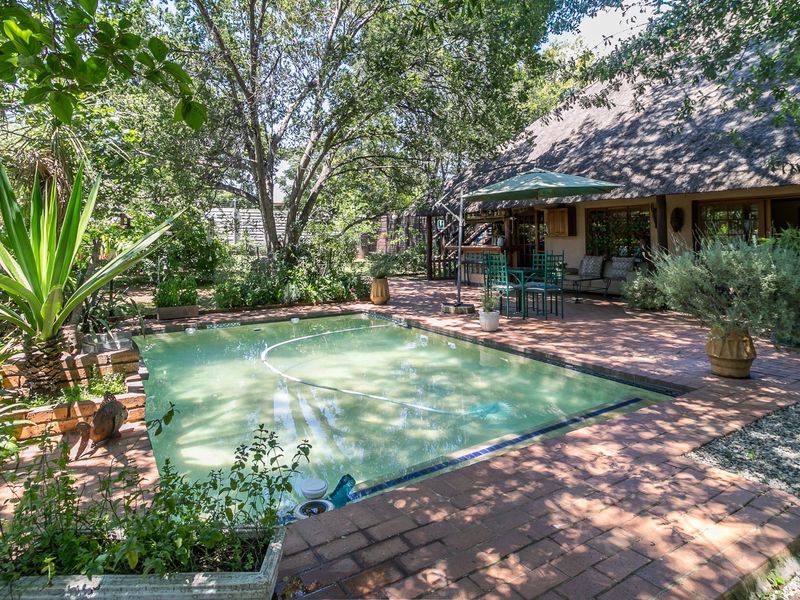 A backyard pool area featuring brick paving, mature garden landscaping, and a house with a traditional thatched roof.