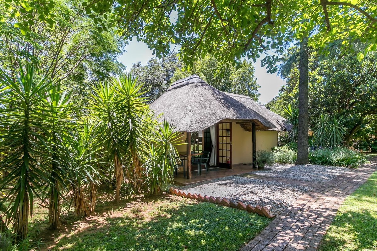 A charming cottage with a traditional thatch roof, featuring a covered patio area set within a lush, mature garden with brick paved pathways.