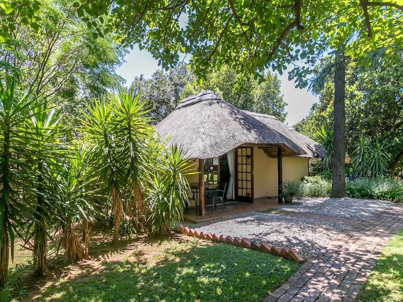 A charming cottage with a traditional thatch roof, featuring a covered patio area set within a lush, mature garden with brick paved pathways.