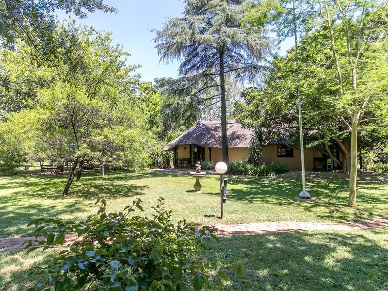 A lush, well-maintained garden featuring mature trees, a manicured lawn, and a brick pathway leading toward a charming thatched-roof dwelling.