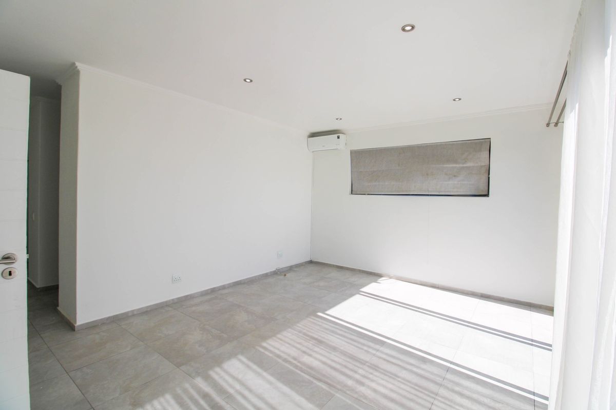 A bright, empty bedroom featuring neutral tiled flooring, recessed downlighting, and a wall-mounted air conditioning unit.