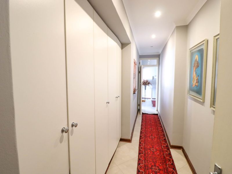 A well-maintained hallway featuring extensive built-in storage cupboards, neutral tiled flooring, and recessed downlighting, leading towards an adjacent room.
