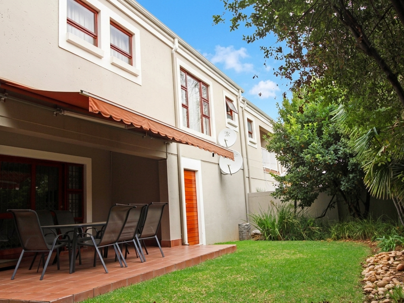 Rear view of the townhouse featuring a covered tiled patio with a retractable awning, overlooking a private landscaped garden and lawn.