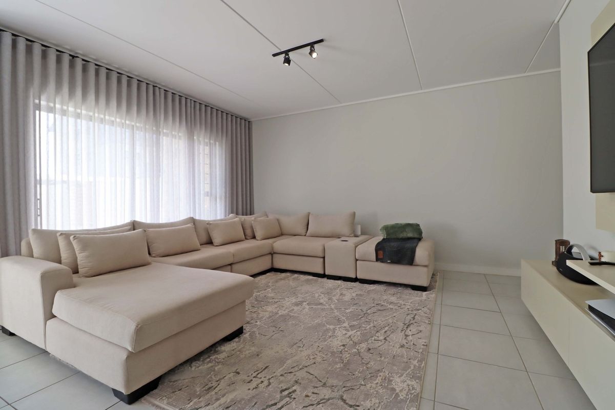 A bright and modern living room featuring neutral tiled flooring, track lighting, and large windows with floor-to-ceiling curtain tracks.