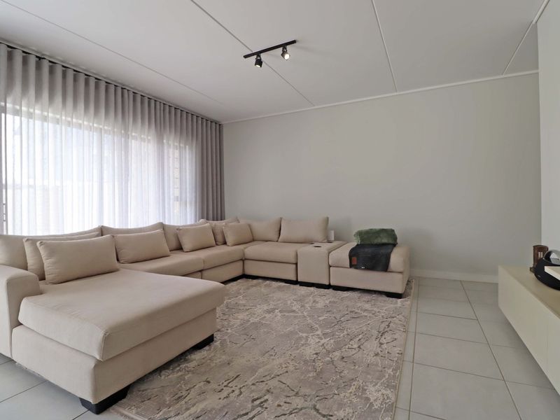 A bright and modern living room featuring neutral tiled flooring, track lighting, and large windows with floor-to-ceiling curtain tracks.