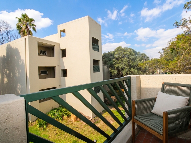 A private balcony featuring a metal balustrade and tiled flooring, overlooking the apartment complex grounds and surrounding greenery.