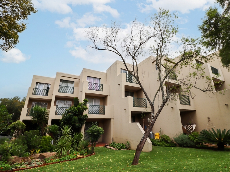A rear view of a multi-story apartment complex featuring private balconies, sliding glass doors, and a well-maintained landscaped garden area.