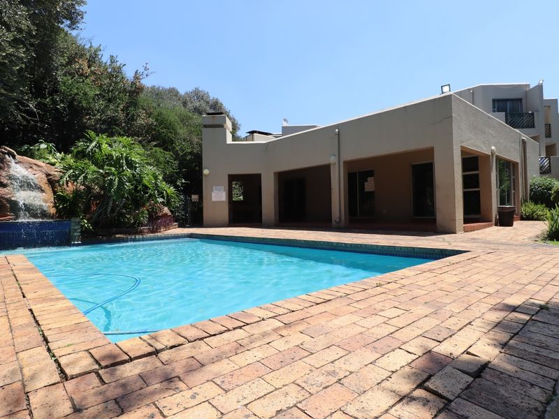 Outdoor pool area featuring a rock waterfall, brick paved surround, and a covered patio space within a landscaped setting.