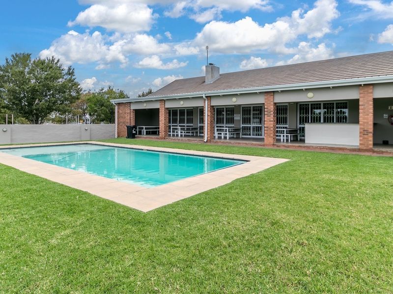 A well-maintained communal pool area featuring a clear swimming pool, paved surround, manicured lawn, and a covered patio with brick structural pillars.