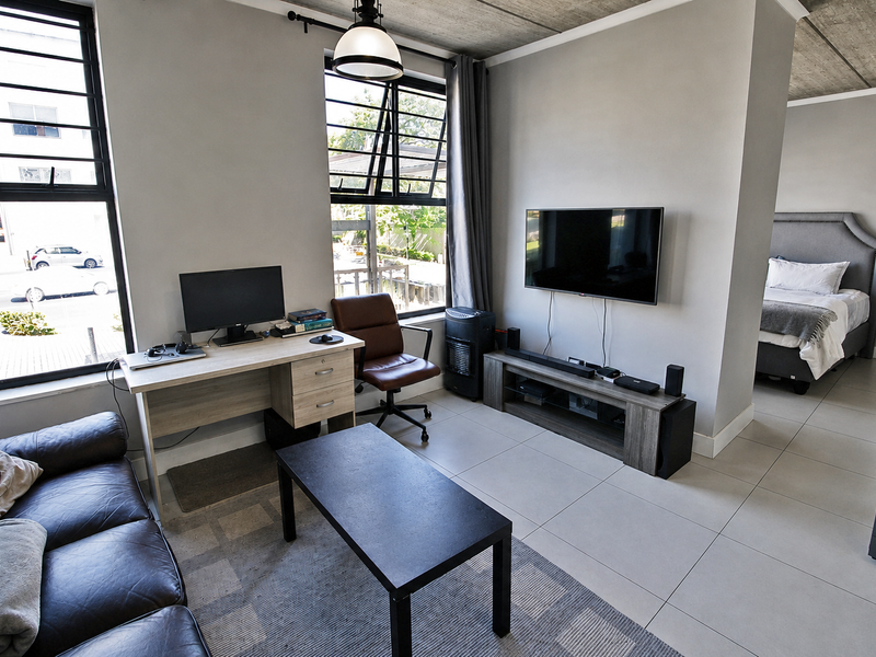 Open-plan living area featuring modern tiled flooring, exposed concrete ceilings, and large windows providing ample natural light, with an adjacent bedroom space.
