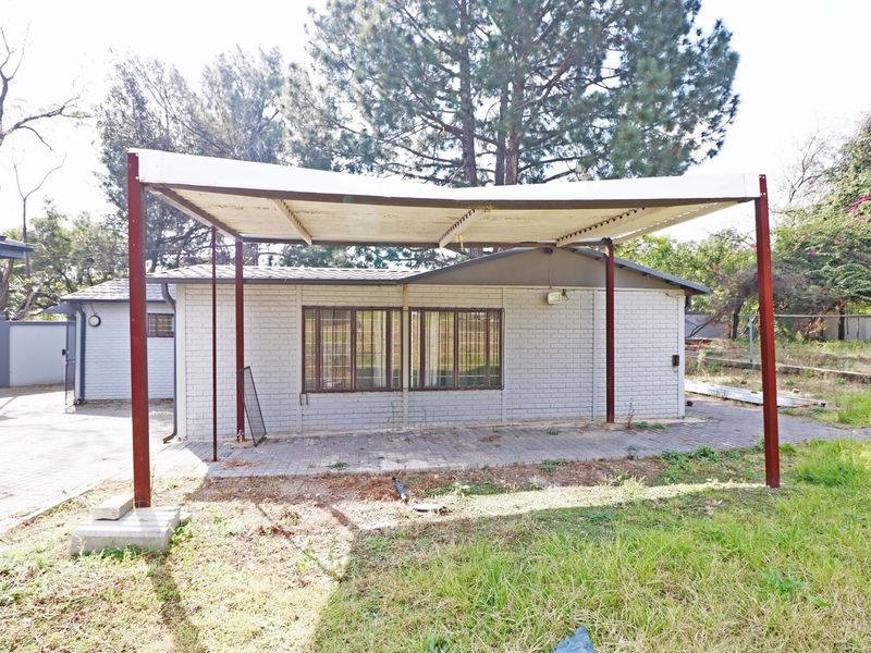 A covered patio area featuring a metal carport structure, paved flooring, and security bars on the windows, set within a residential garden space.