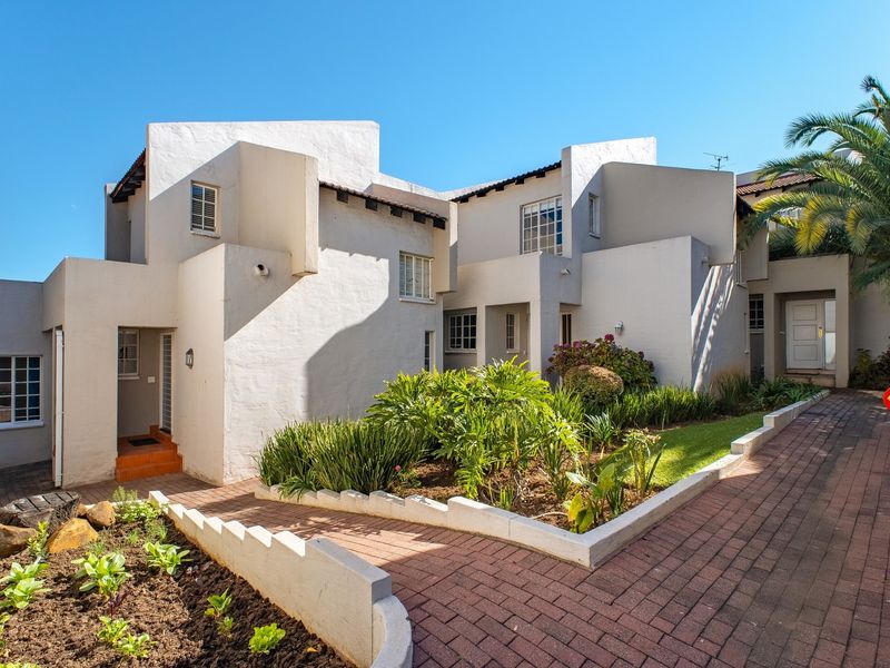 Front elevation of a townhouse complex featuring a paved walkway, landscaped garden beds, and white stucco exterior walls.