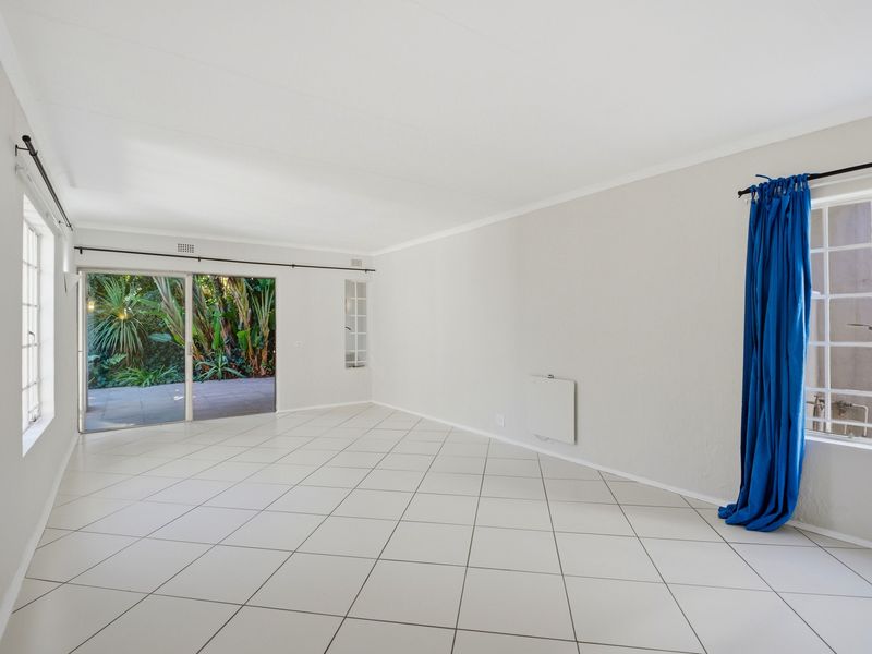 A bright, empty living room featuring neutral tiled flooring, a wall-mounted heater, and sliding glass doors providing direct access to the rear patio and garden.