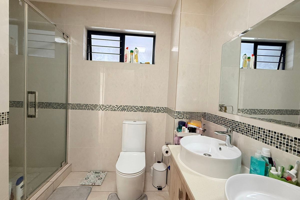 A well-maintained bathroom featuring a glass shower enclosure, dual vanity basins with storage cabinetry, and neutral wall tiling with a decorative mosaic border.