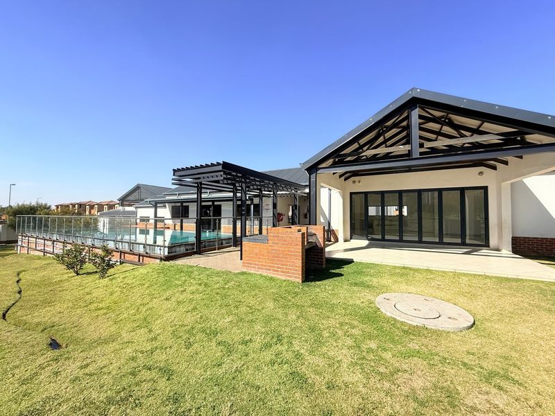 Rear exterior view featuring a swimming pool with glass balustrades, a brick built-in braai, a covered pergola, and a spacious tiled patio area.