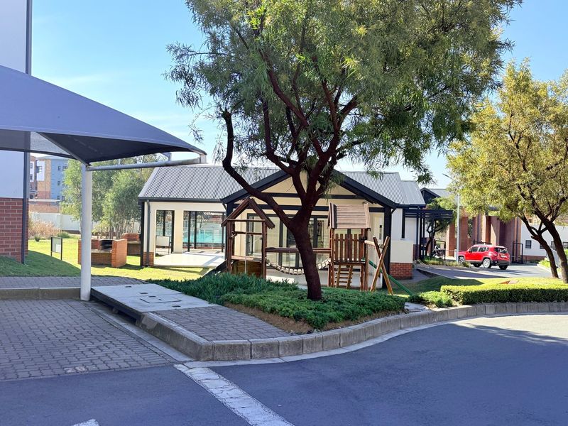 Exterior view of a communal clubhouse facility featuring a brick braai area, shade sail, and landscaped garden within the apartment complex.