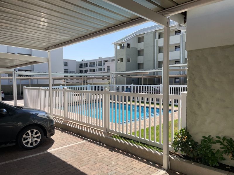 A view of the communal swimming pool area featuring a paved surround, white safety fencing, and adjacent covered parking bays.